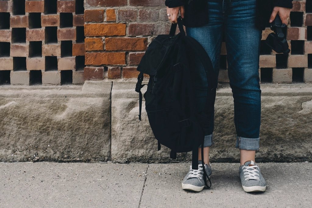 Black backpack held by its handle next to cuffed jeans against a brick wall