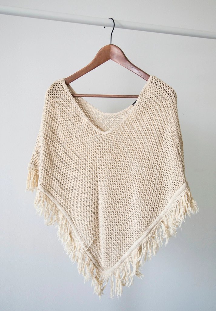 Cream open-knit fringed poncho on a wooden hanger against a white wall