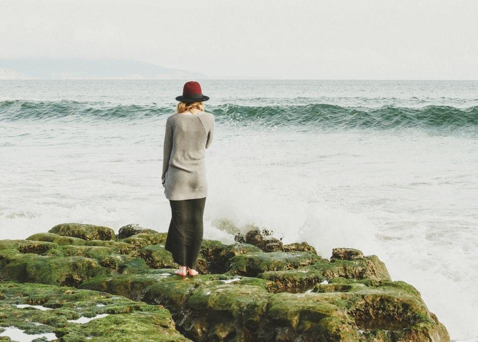 Model in a grey knit and hat standing on rocks by the sea