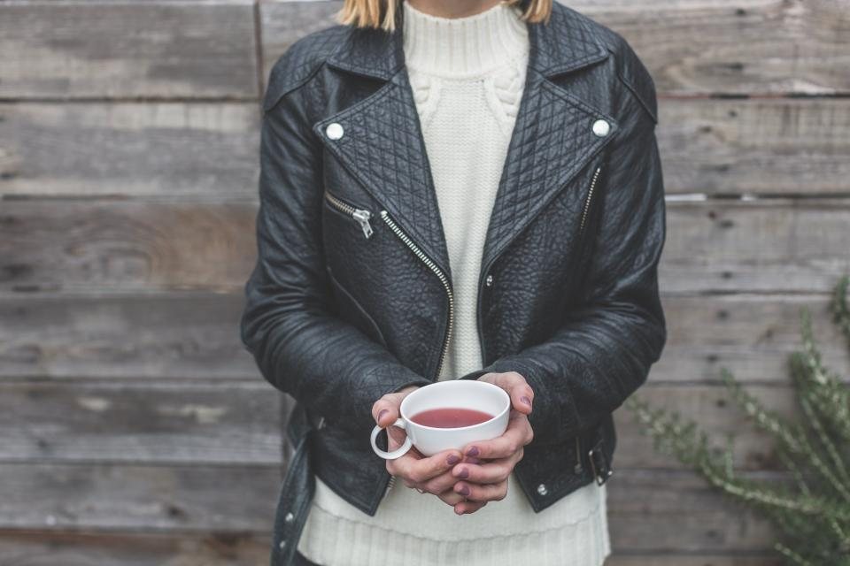 Woman in a black leather biker jacket and cream knit holding a cup of tea, face out of frame