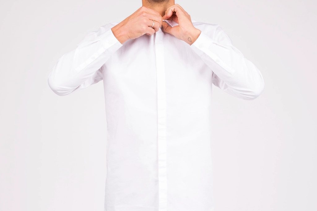 Man buttoning a crisp white shirt against a light grey studio wall, face out of frame
