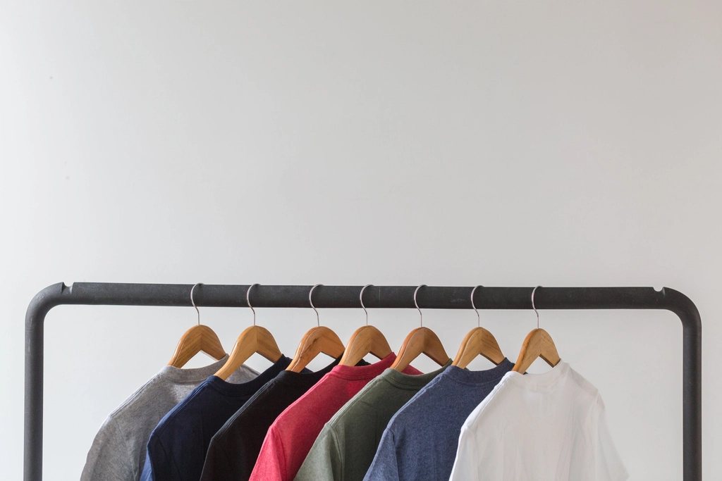 Seven plain tees on wooden hangers along a black steel rail against a white wall