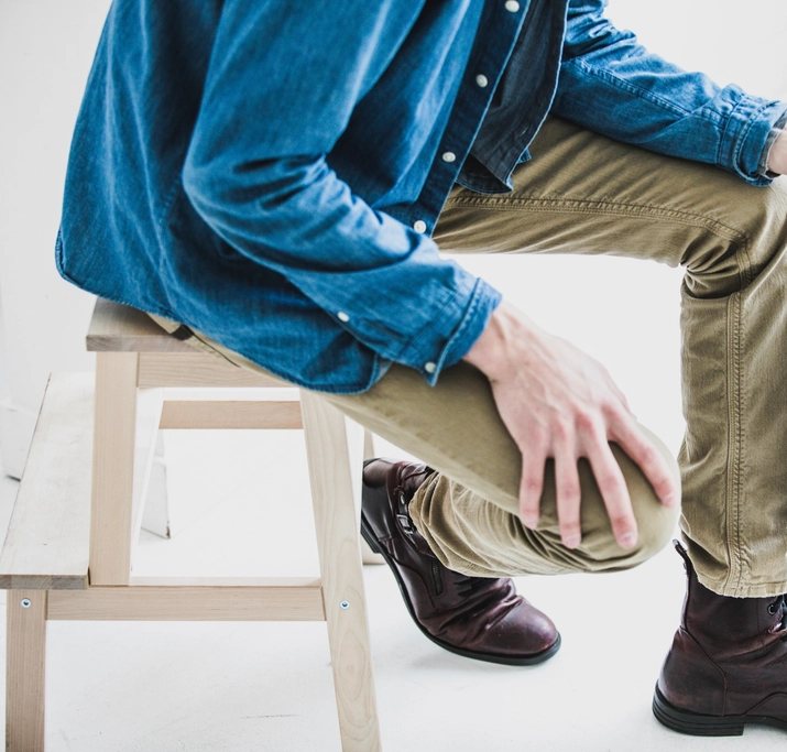 Seated man in khaki trousers, a denim shirt and burgundy leather boots on a wooden stool