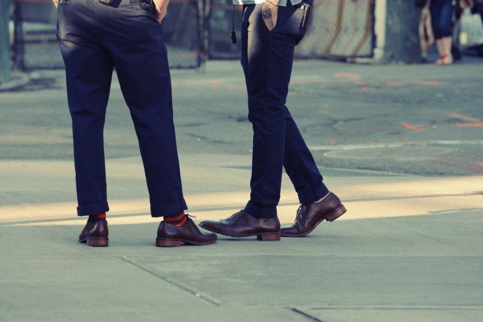 Two people in cuffed navy trousers and brown leather shoes on a city pavement